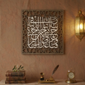 An ornate square wall art piece with white Arabic calligraphy set in a carved brown frame, displayed on a textured terracotta wall above a carved console table with books and a brass lamp.